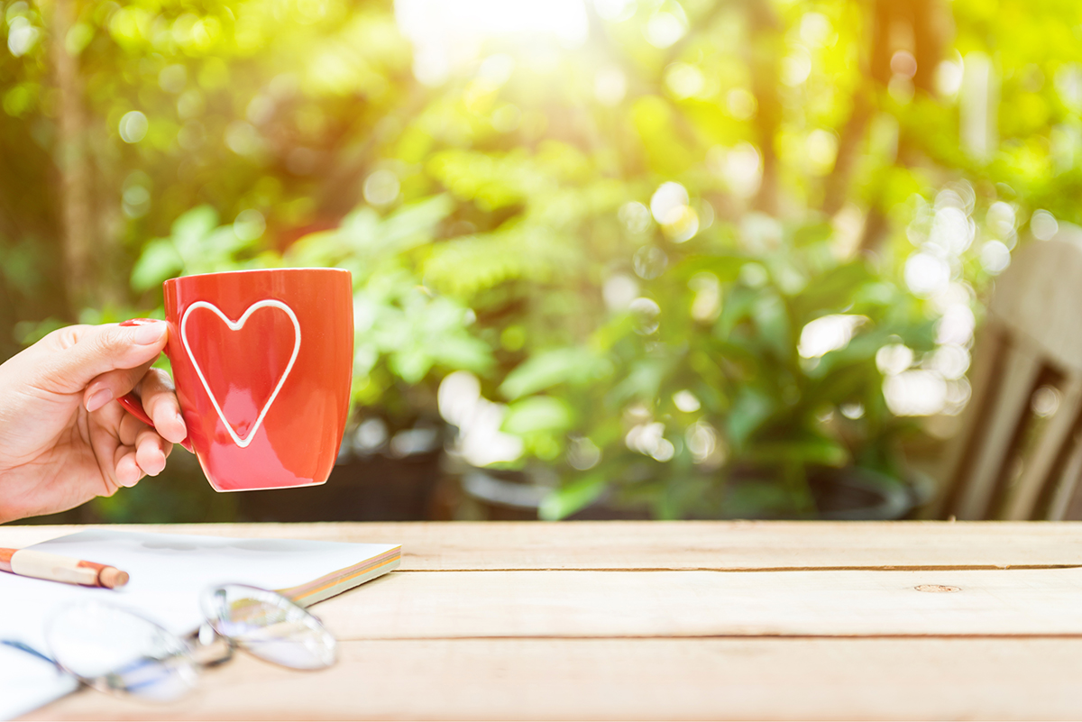 Käsi pitelee punaista, sydämen kuvalla varustettua mukia puutarhassa.