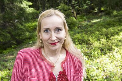 Hymyilevä Kaisa Hartikainen punaisessa puserossa ja taustalla vihreää metsää.