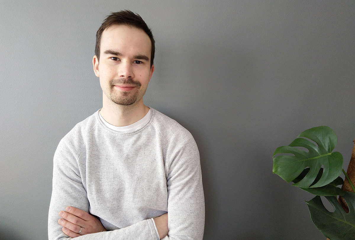 Hymyilevä nuorimies valkoisessa paidassa, taustalla harmaa seinä ja viherkasvi.