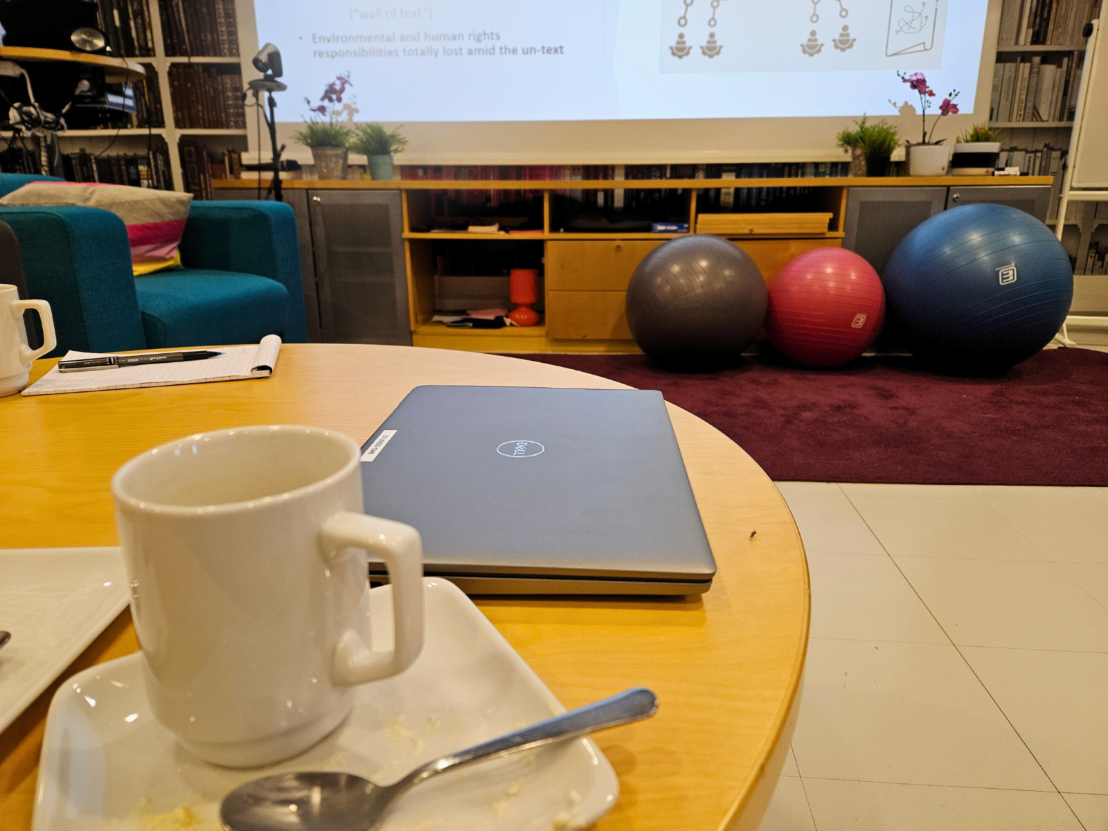 Kuva seminaarihuoneesta. Etualalla näkyy pöytää, jolla on kahvikuppeja, tyhjä kakkulautanen sekä läppäri. Taustalla näkyy värikkäitä sisustuselementtejä sekä hieman valkokankaan alareunaa ja sille heijastettua esitelmää.