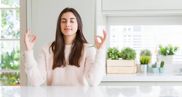 Tummatukkainen nuori nainen valoisassa keittiössä meditoimassa.