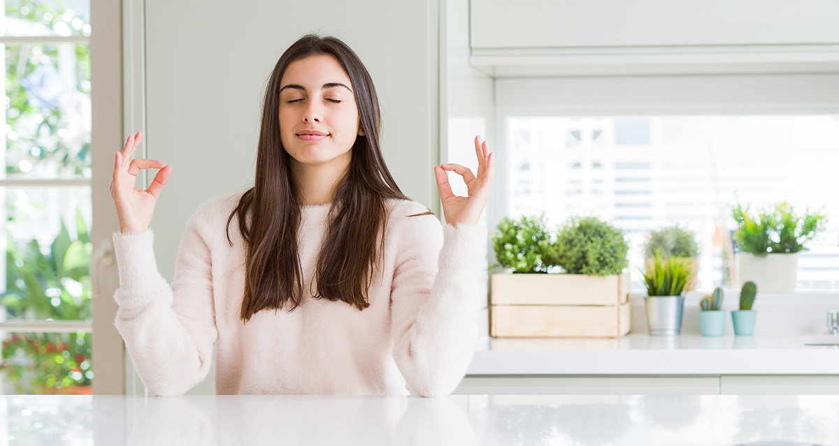 Tummatukkainen nuori nainen valoisassa keittiössä meditoimassa.