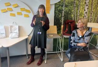 Woman standing and explaining, another woman listening to her. 