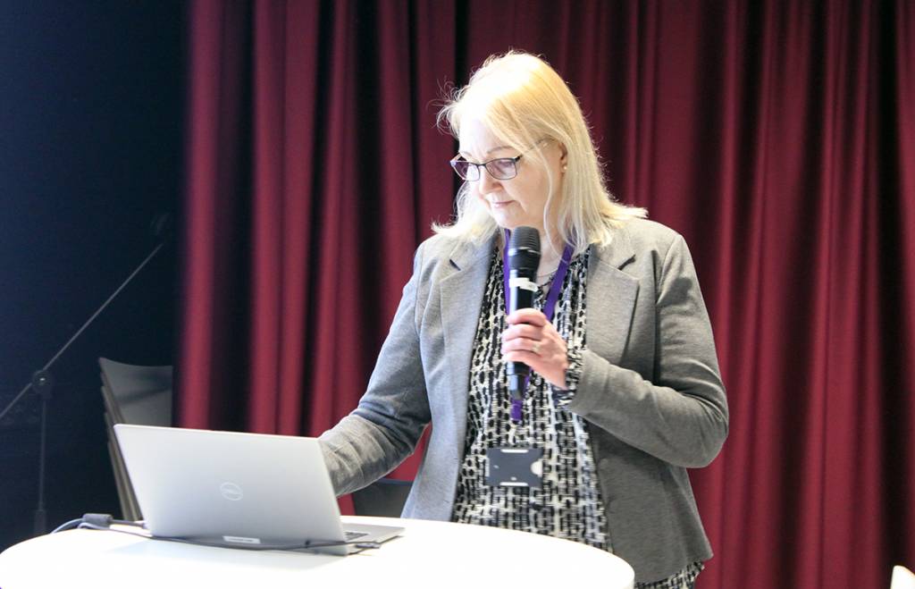 Vaaleatukkainen nainen mikrofoni kädessä läppärin ääressä.