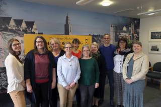 A group of 10 people standing and smiling in front of the wall with picture of a college building n n