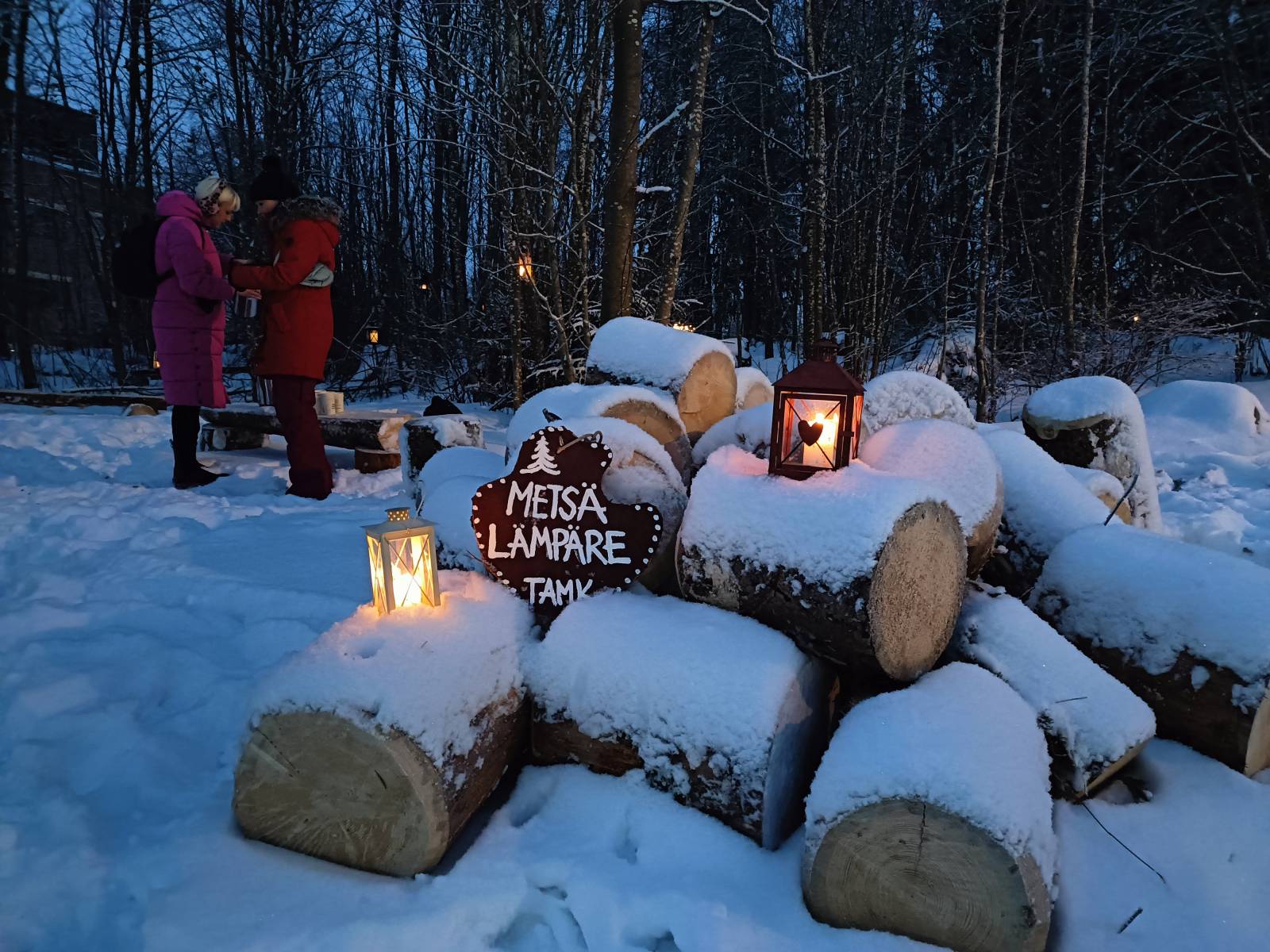 Lumisen puupölkkykasan päällä Metsälämpäre-kyltti ja kaksi lyhtyä