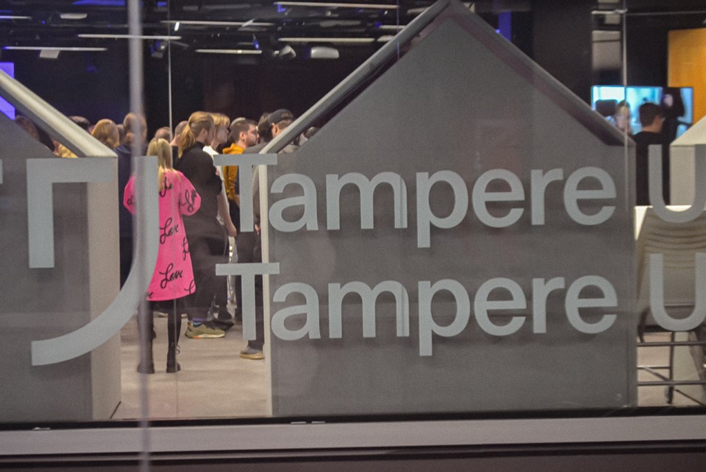 Kuvassa näkyy ikkuna, jossa teksti Tampere. Ikkunan takana näkyy sisätiloissa oleskelevaa väkijoukkoa.