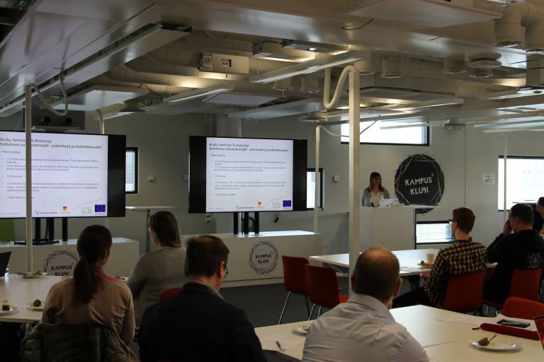 Ihmisiä selin kameraan seminaatilassa. Projektikoordinaattori Johanna Lahti pitää esiltelmää