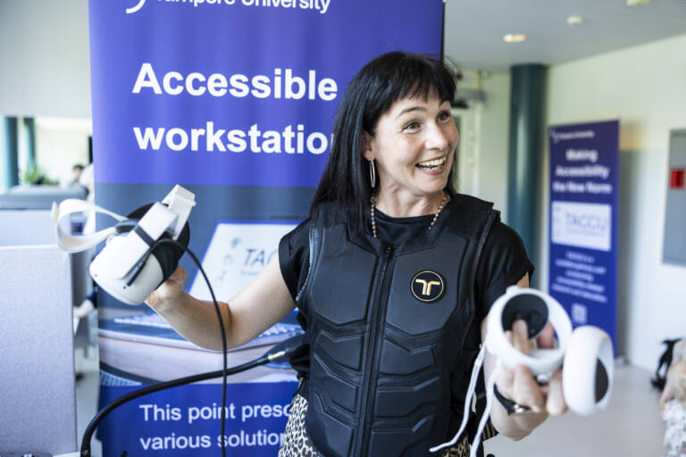 Liisa Mustanoja hymyilee VR-lasit kädessään. Hänellä on yllään tuntopalautetta antava haptinen liivi.