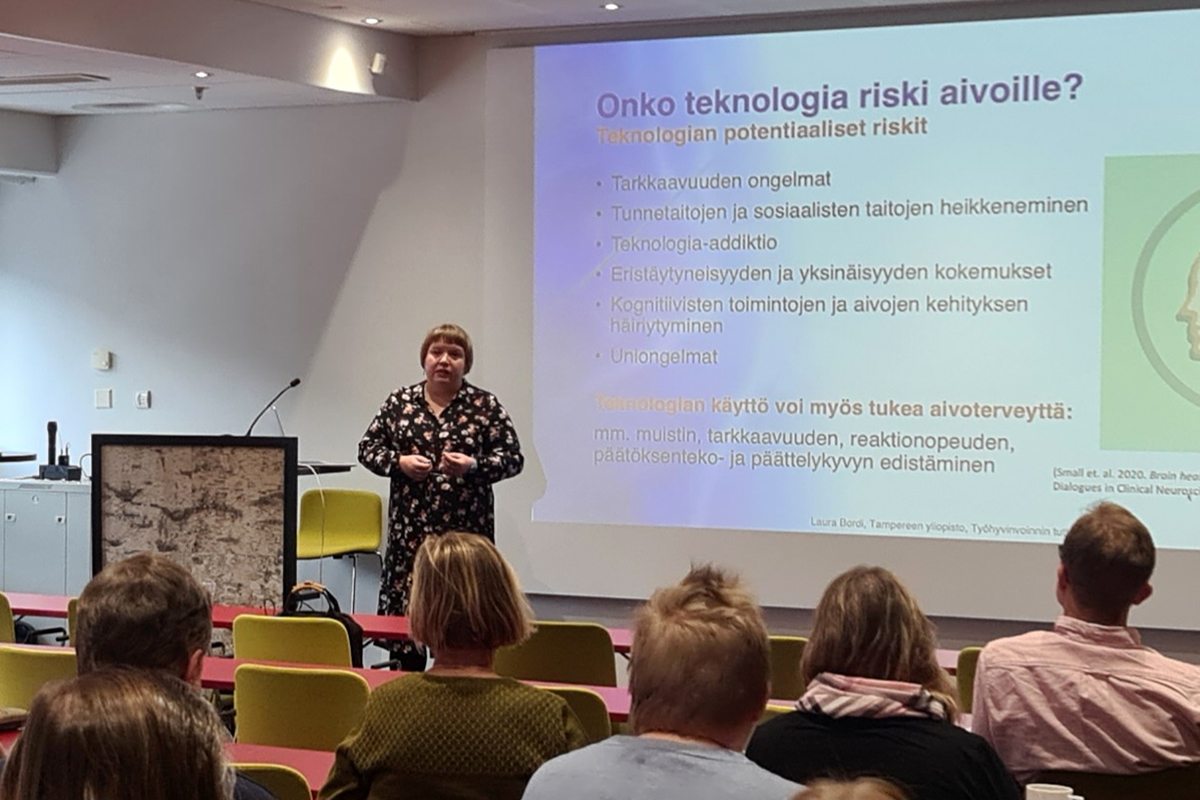 Lyhyttukkainen nainen pitää esitystä, yleisö kuuntelee.