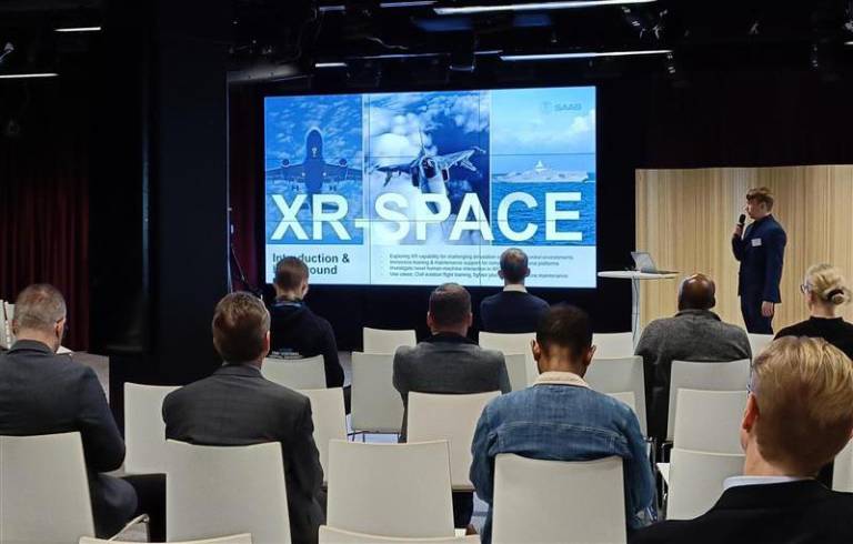 People are sitting in chairs in a room and watching screen. There is a word XR-SPACE on the screen.