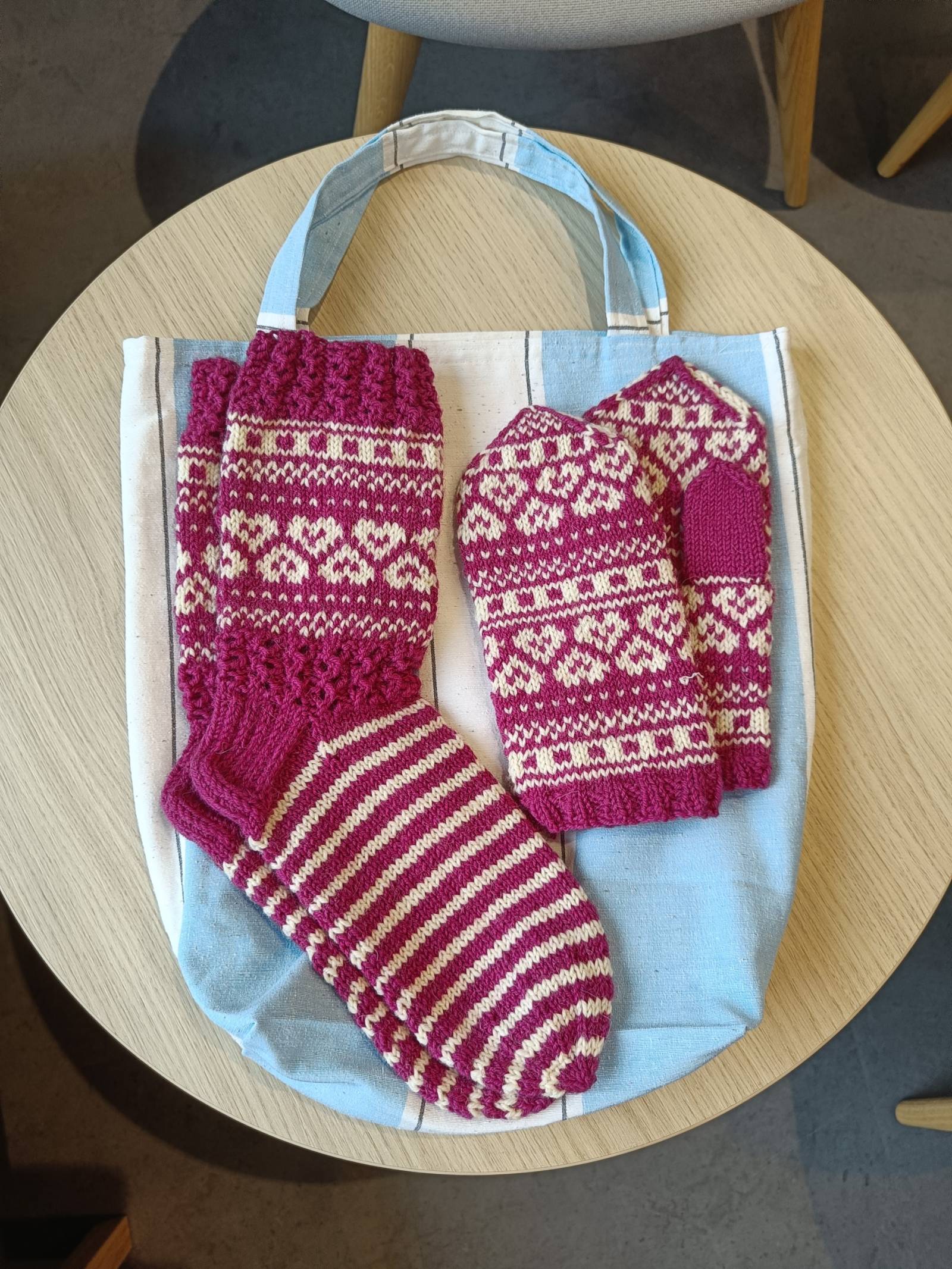 Pöydällä sinisen kassin päällä punavalkoiset kirjoneulesukat ja kirjoneulelapaset.