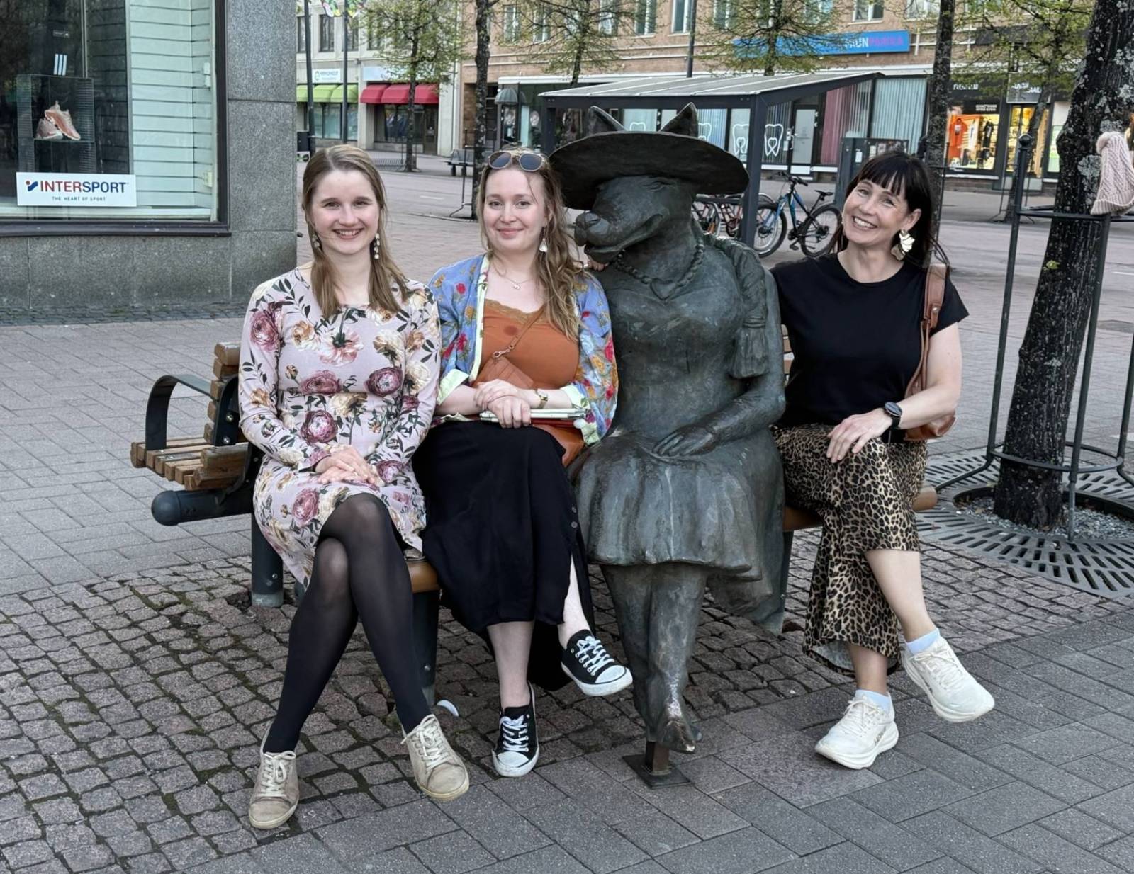 Juuli, Vilma ja Liisa istuvat ulkona penkillä, jolla istuu myös naistenvaatteisiin pukeutunutta sutta esittävä patsas.