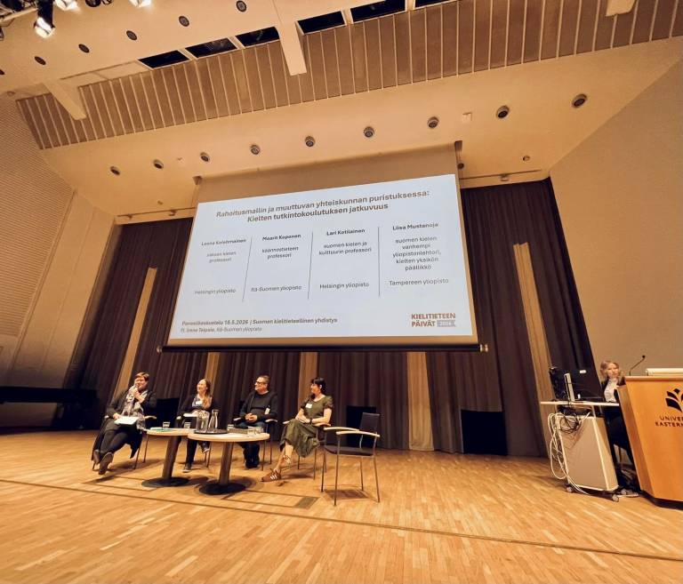 Neljä panelistia istuu pyöreiden pöytien ääressä auditorion lavalla. Paneelin puheenjohtaja seisoo lavalla kauempana tietokoneen takana. Panelistien taakse valkokankaalle on heijastettu teksti, jossa lukee: Rahoitusmallin ja muuttuvan yhteiskunnan puristuksessa: Kielten tutkintokoulutuksen jatkuvuus. Leena Kolehmainen, saksan kielen professori, Helsingin yliopisto. Lari Kotilainen, suomen kielen ja kulttuurin professori, Helsingin yliopisto. Maarit Koponen, käännöstieteen professori, Itä-Suomen yliopisto. Liisa Mustanoja, suomen kielen vanhempi yliopistonlehtori, kielten yksikön päällikkö, Tampereen yliopisto. Pj. Irene Taipale, Itä-Suomen yliopisto.