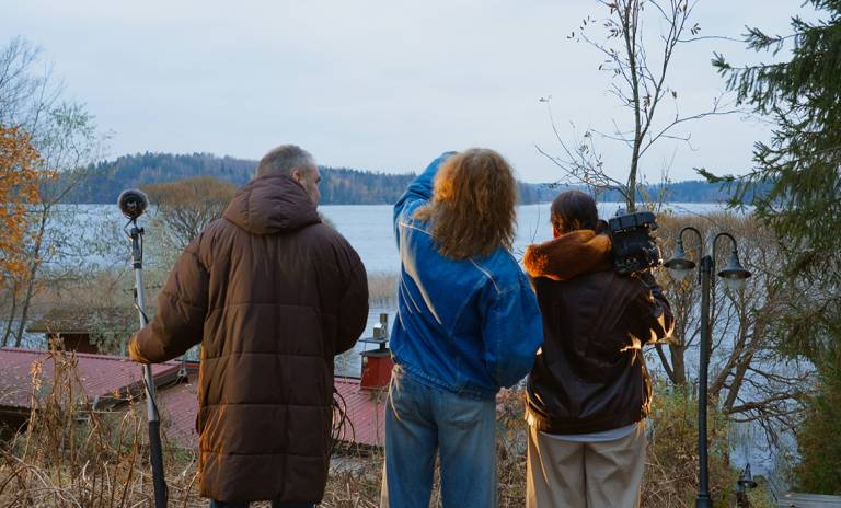 Kolme henkilöä järven rannalla käsissään kamera ja mikrofoni.