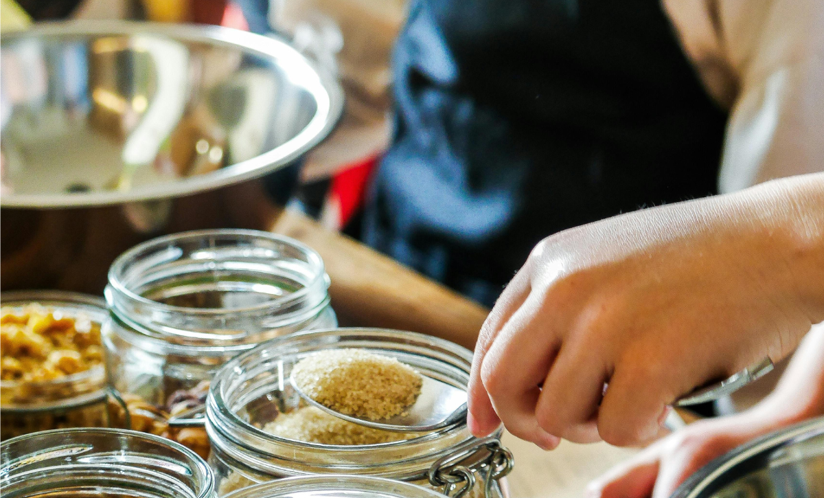 Ihminen otta lusikallisen sokeria purkista ruoanlaittotilanteessa