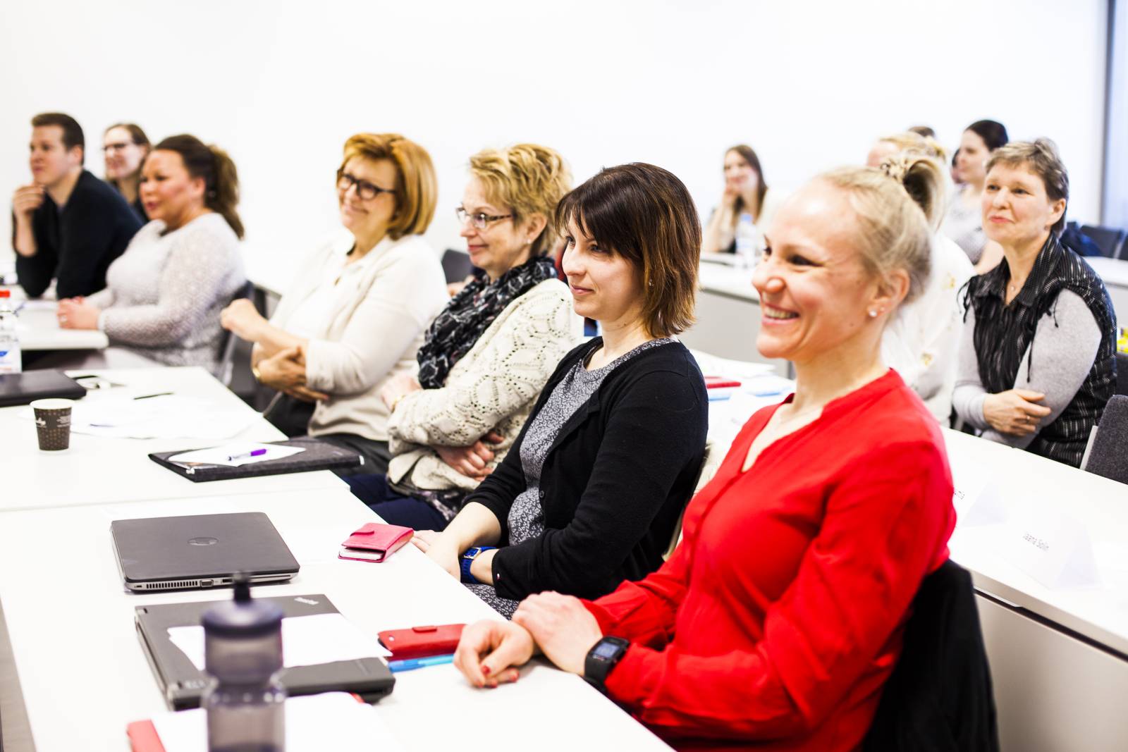 Luentosalissa istuu ihmisiä kuuntelemassa opetusta. He hymyilevät ja vaikuttavat innostuneilta.