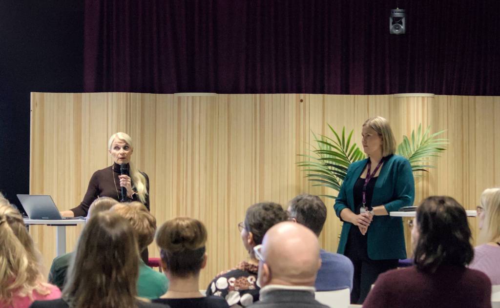 Kaksi esiintyjää seisoo lavalla puhumassa yleisölle, taustalla puinen seinä.