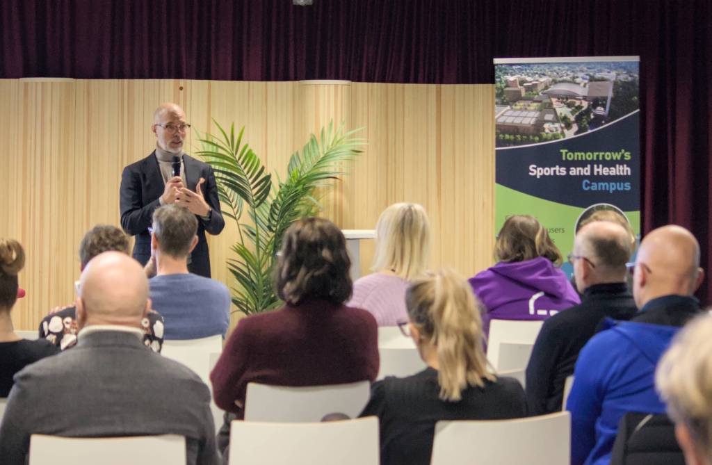 Puhuja seisoo lavalla yleisön edessä, taustalla puinen seinä ja Tomorrow’s Sports and Health Campus -roll-up.