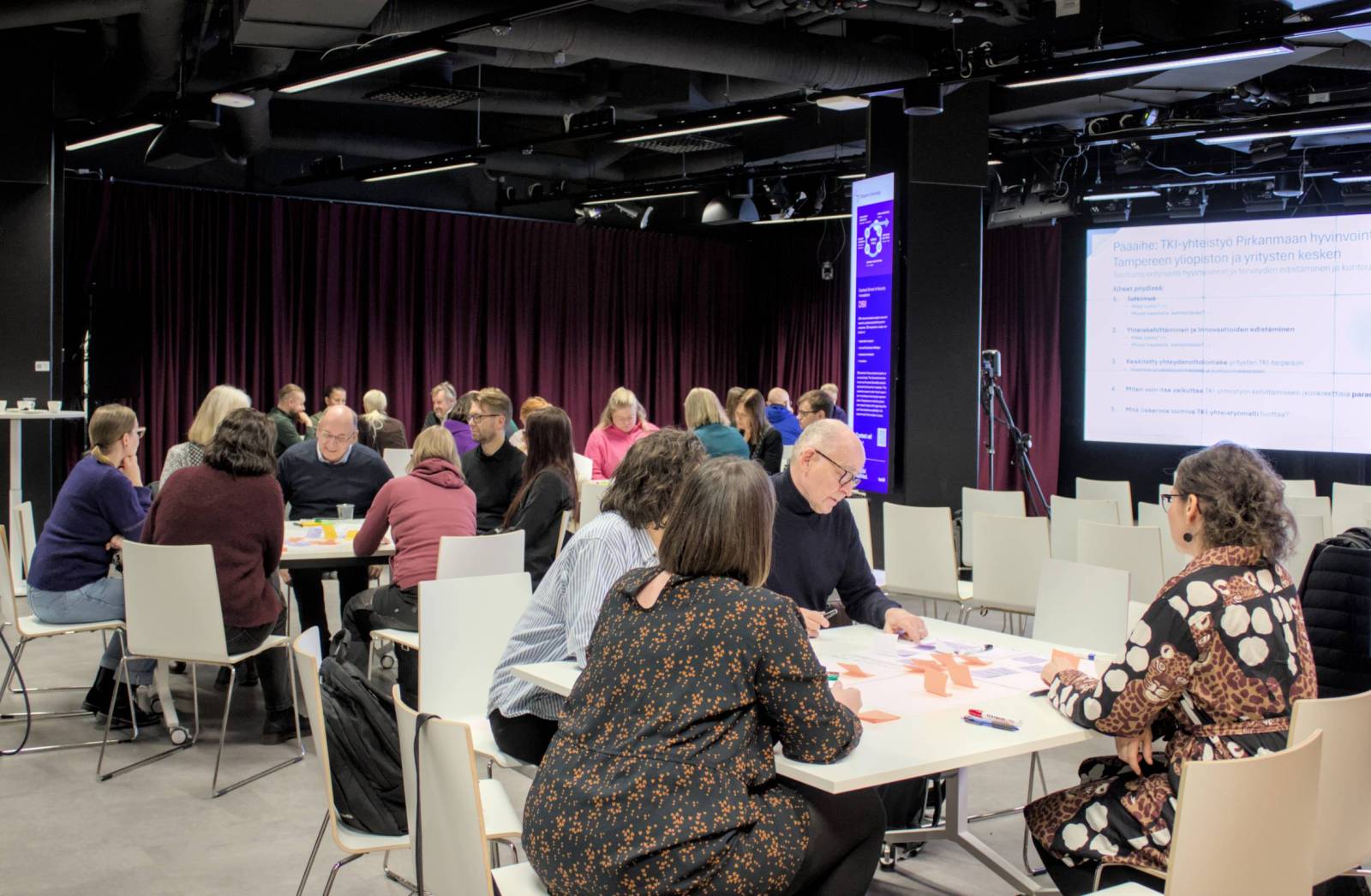 Ryhmä ihmisiä istuu pöytien ääressä modernissa neuvottelutilassa osallistuen työpajaan. Taustalla on suuri näyttö, jossa näkyy esitysdia, sekä tummat verhot ja valaistut infonäytöt seinillä.