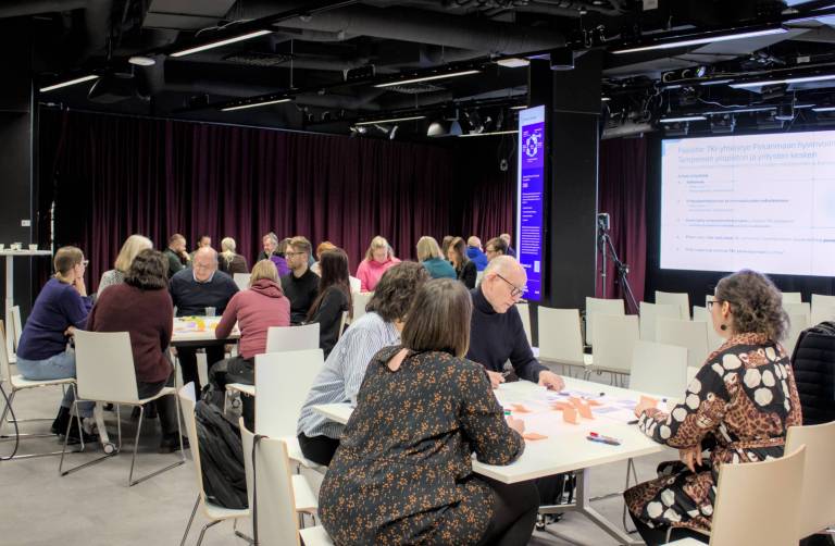 Ryhmä ihmisiä istuu pöytien ääressä modernissa neuvottelutilassa osallistuen työpajaan. Taustalla on suuri näyttö, jossa näkyy esitysdia, sekä tummat verhot ja valaistut infonäytöt seinillä.