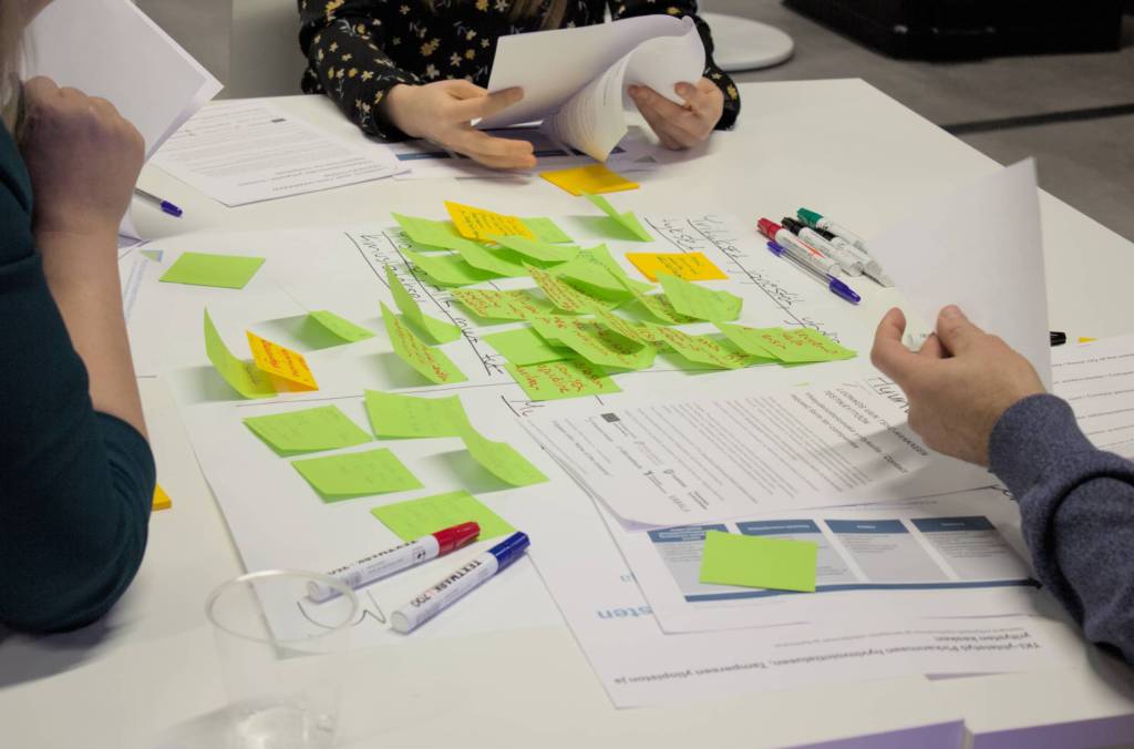 Työpajapöytä, jossa papereita, värikkäitä muistilappuja ja tusseja, osallistujat tekevät muistiinpanoja.