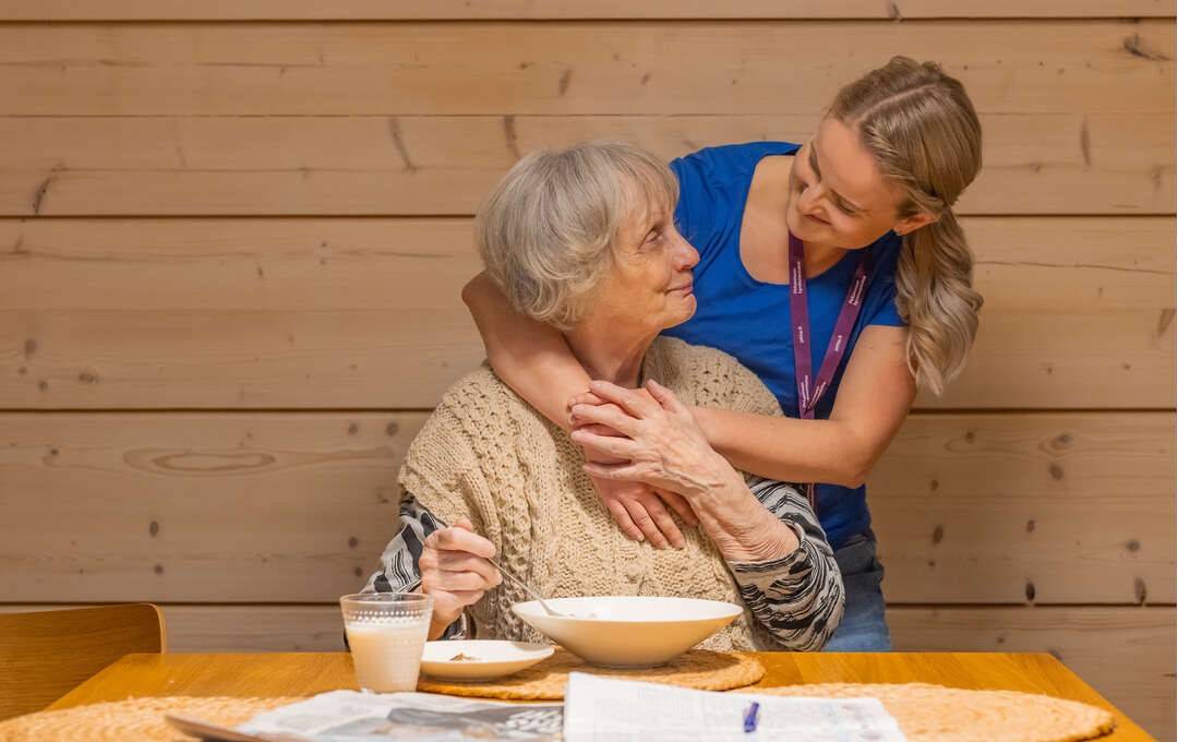 Ikääntynyt nainen istuu ruokapöydän ääressä, edessään lautanen, lasi ja sanomalehti. Taustalla sairaanhoitaja kumartuu halaamaan olkapäiden ympäriltä.