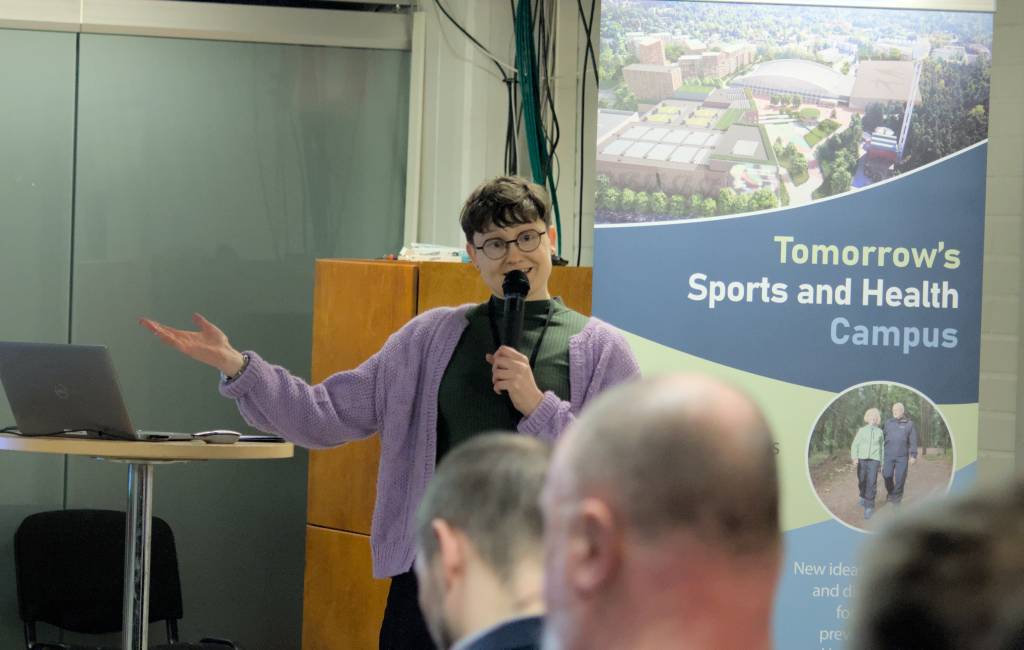 Puheenpitäjä esittelee aihettaan mikrofonin kanssa yleisölle, taustalla “Tomorrow’s Sports and Health Campus” -esittelybanneri.