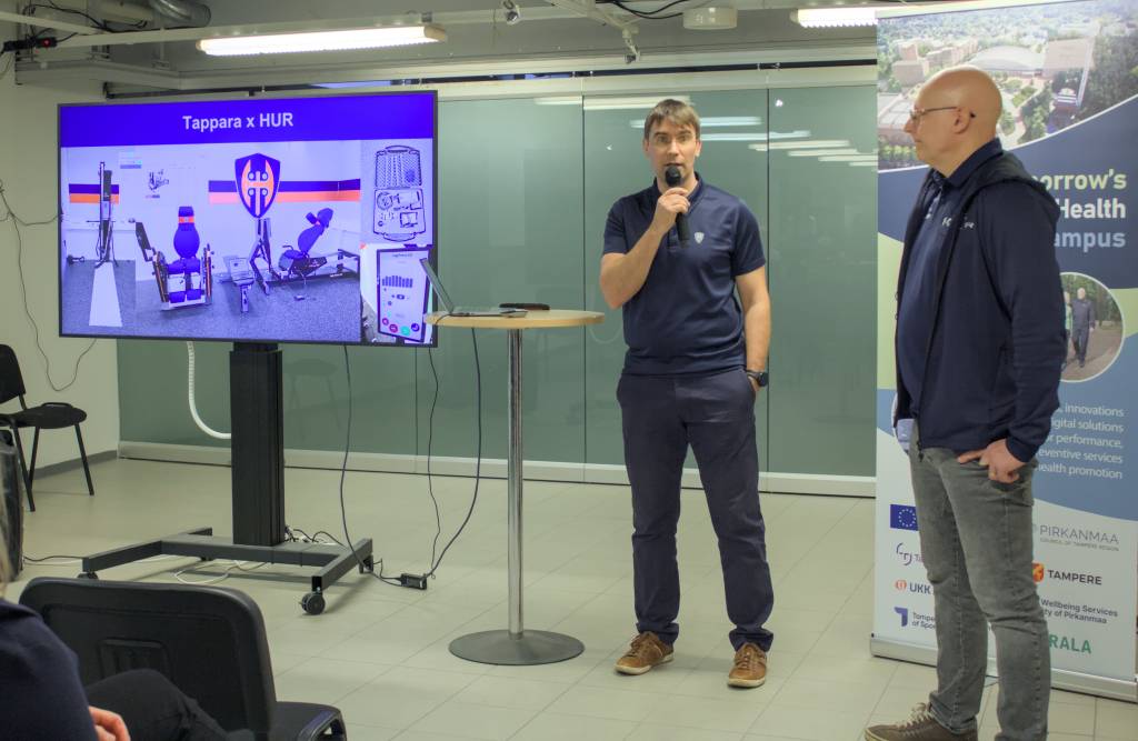 Kaksi esiintyjää esittelee “Tappara x HUR” -yhteistyötä. Taustalla näyttö, jossa näkyy kuntosalilaitteita.
