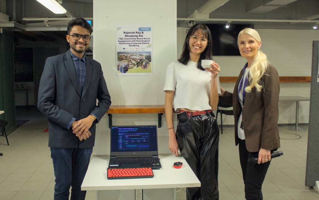 Kolme henkilöä poseeraa esittelypisteellä pöydän ja kannettavan tietokoneen edessä, taustalla tapahtumamainos.