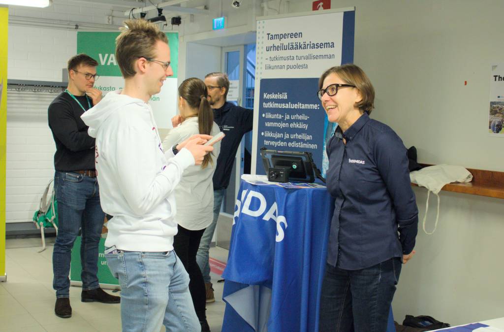 Kaksi henkilöä keskustelee verkostoitumisosuudessa, taustalla Tampereen urheilulääkäriaseman esittelypiste.