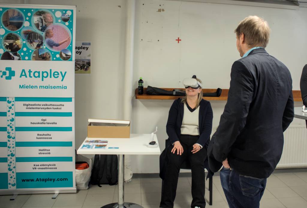 Henkilö kokeilee VR-laseja istuen esittelypisteellä, vieressä Atapley-yrityksen esittelyroll-up.