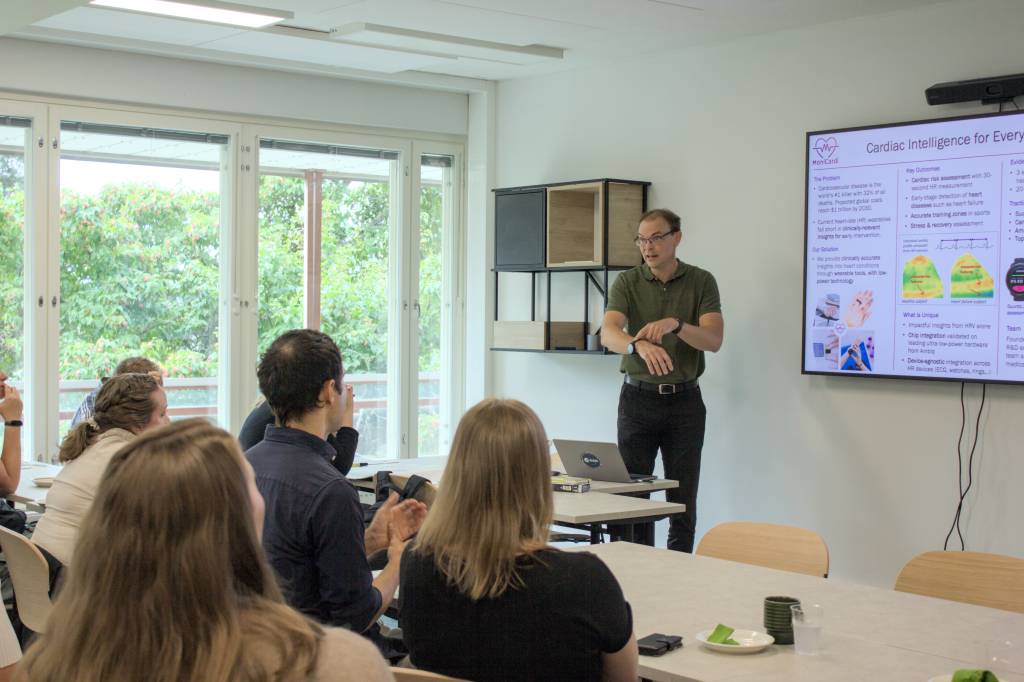 Mies pitää esitystä ja osoittaa älykelloaan. Etualalla yleisöä istumassa tuoleilla.