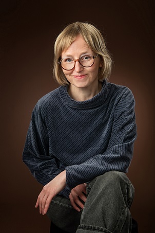 Kuvassa on hankkeen tutkija Katariina Mäkinen. Mäkinen katsoo kameraan hymyillen. Hän nojaa toisella kyynärpäällään polveensa.