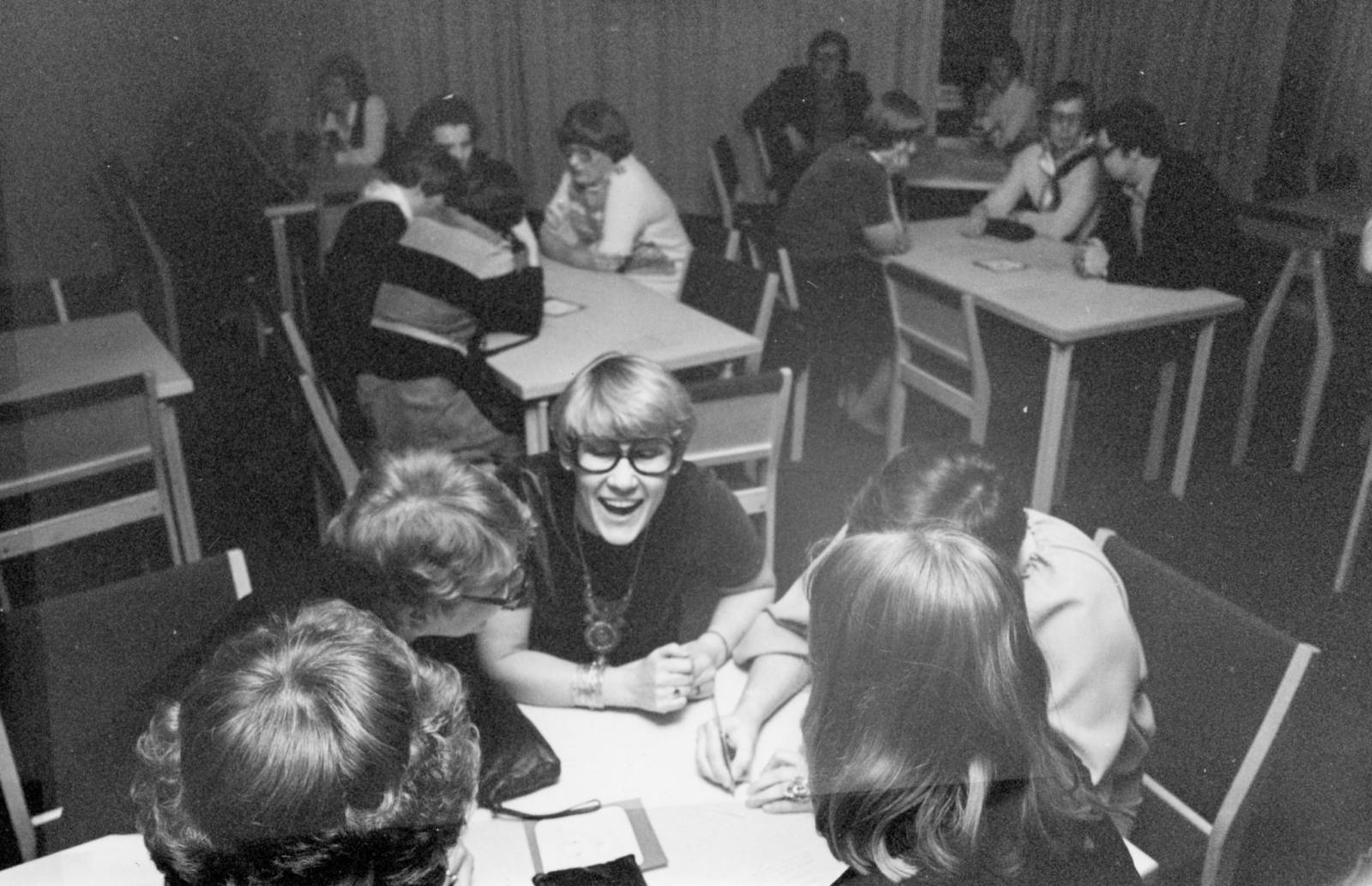 Mustavalkoisessa kuvassa joukko aikuisia ihmisiä istuu pienissä ryhmissä kolmen pöydän ympärillä. Keskellä kuvaa on naurava, vaalea silmälasipäinen ihminen.