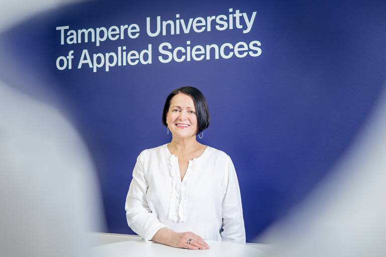 Hymyilevä Mirva Kolonen, taustalla Tampereen ammattikorkeakoulun englanninkielinen logo.