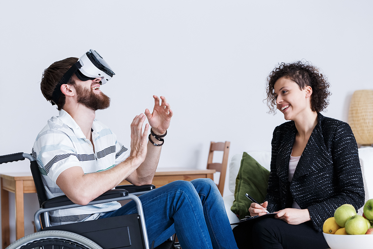 Pyörätuolissa istuva mies VR-lasit päässä ja hänen vieressään nuori nainen.
