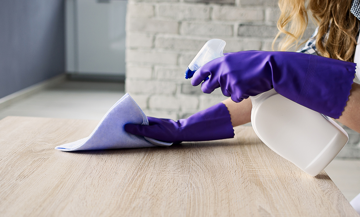 Violetein siivoushansikkain varustetut kädet pyyhkivät vaaleata puupintaa valkoisella siivousliinalla ja suihkaisevat pesuainetta pullosta.