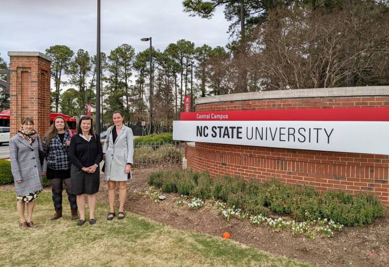 Neljä naista poseeraamassa North-Carolina State Universityn ulkokyltin vieressä.