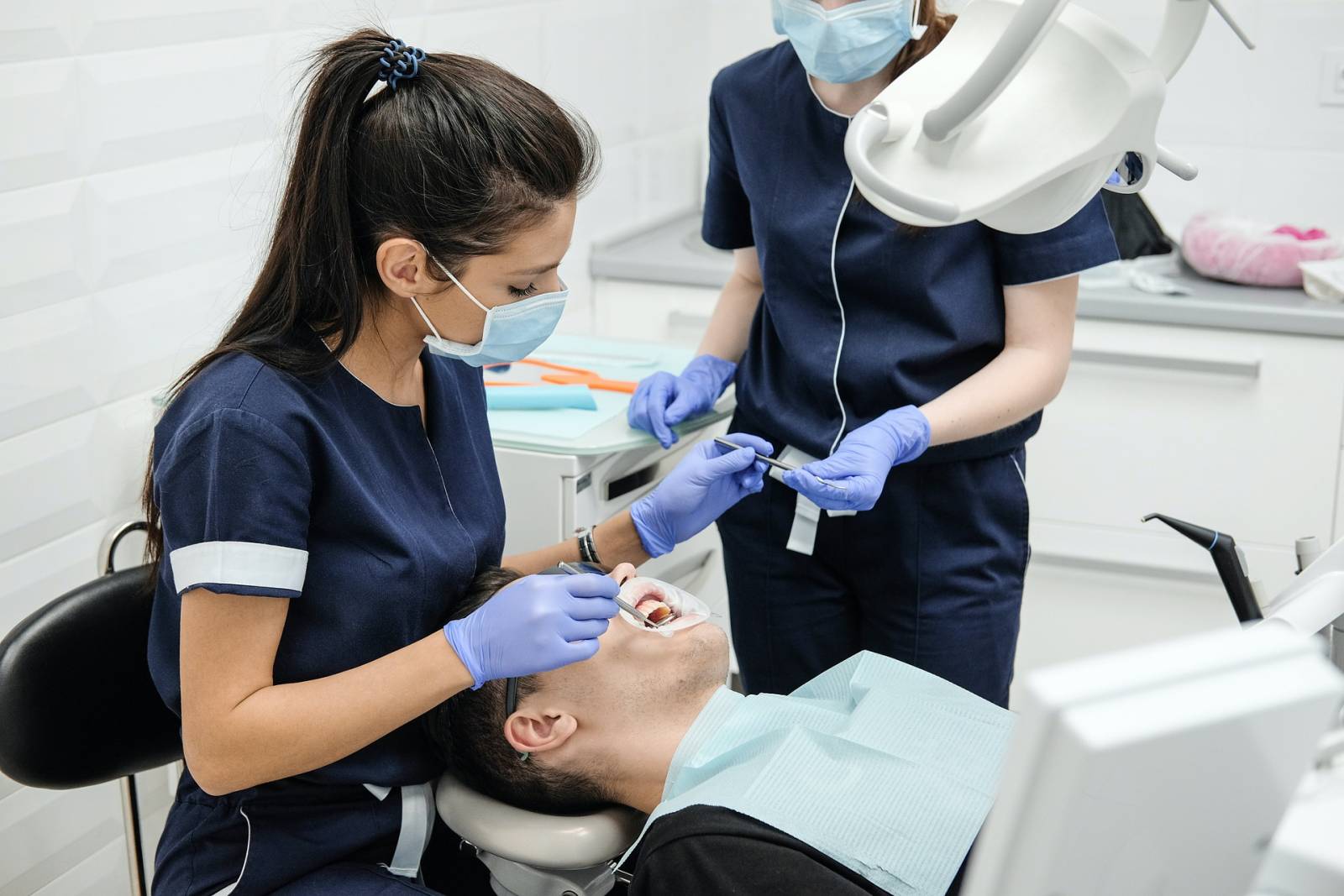A dental nurse/dentist inspecting a lying patients mouth while another dental nures/dentist is handing an instrument to them.