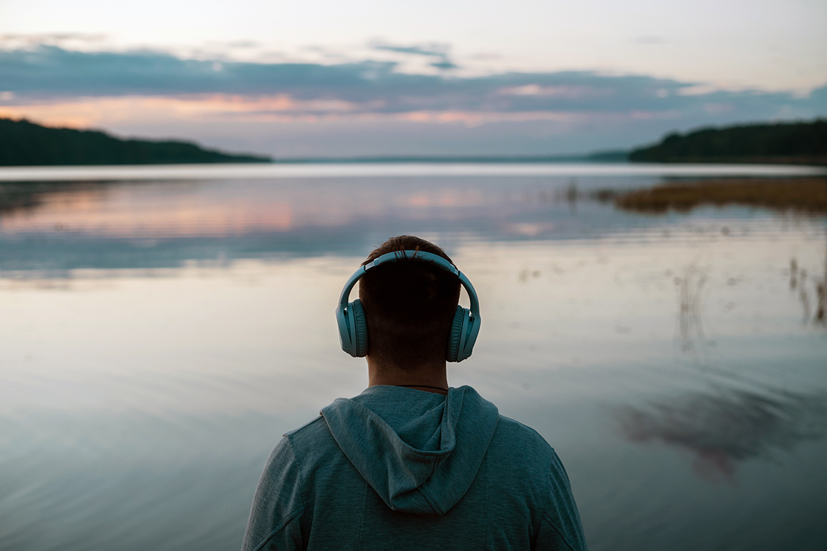 Mies kuuntelee kuulokkeista musiikkia järven rannalla auringon laskiessa.
