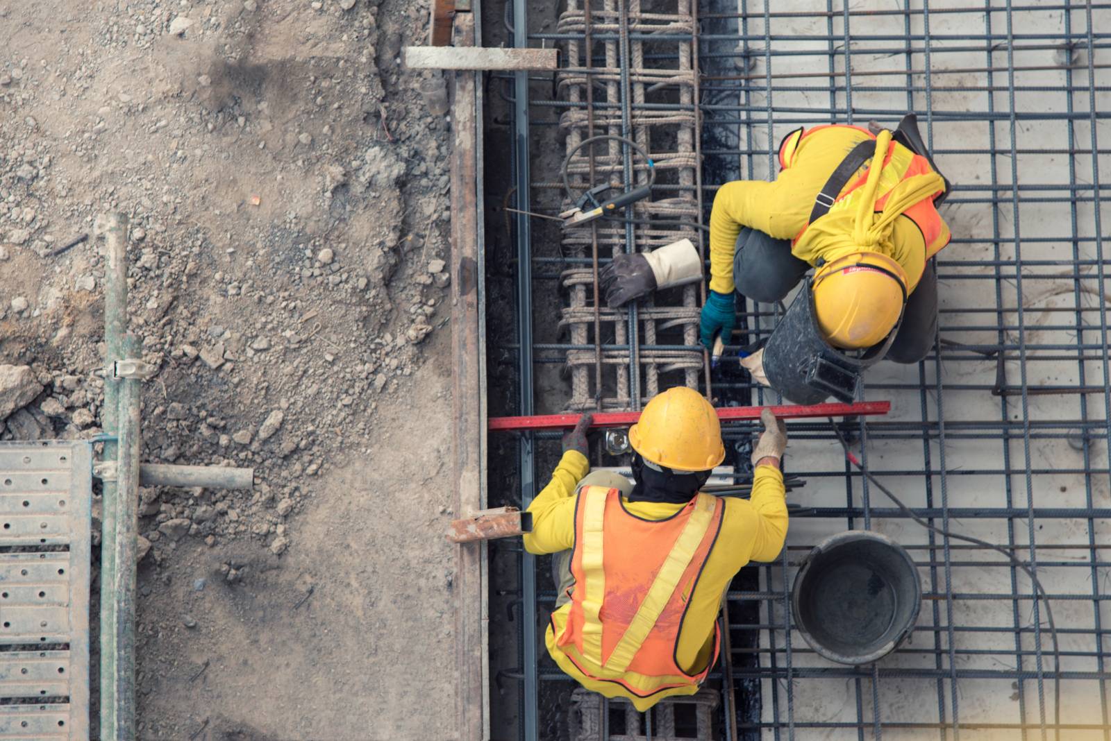 Kuvattu ylhäältä päin, kaksi rakennustyöntekijää työskentelemässä rakennustyömaalla huomioliiveissä.