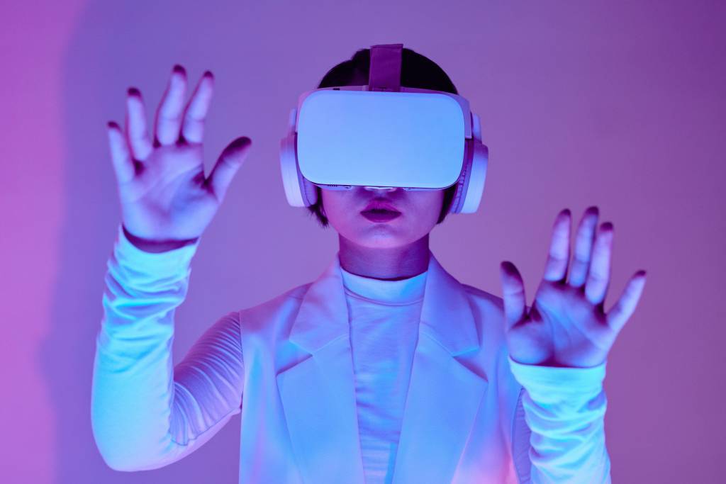 Lähikuva valkoisiin vaatteisiin pujeutuneesta naisesta, jolla silmillään VR-lasit ja korvillaan kuulokkeet. Kädet pystyssä kuvaajaan päin. Futuristinen kuva.