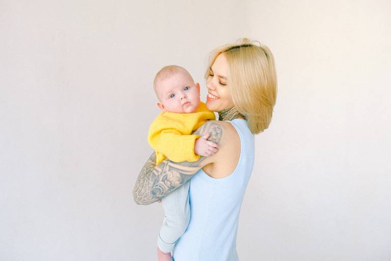 Tatuoitu äiti pitää keltaiseen villapaitaan puettua vauvaa sylissä