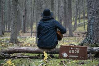 Kuvituskuva. Henkilö istuu metsässä puunrungolla ja soittaa kitaraa.
