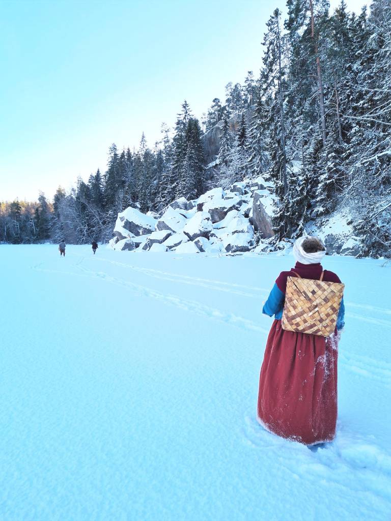 1600-luvun naispuolinen elävöittäjä kävelee talvimaisemassa järven jäällä. Hänellä on punainen hame ja tuohikontti selässään.