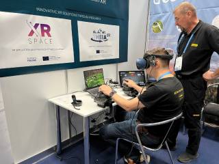 A man wearing XR glasses is sitting by a table using controllers with both of his hands and watching a laptop screen. Next to him there is another man standing and watching the same screen. On the wall in front of the table there is a big poster with a XR-SPACE project logo on it.