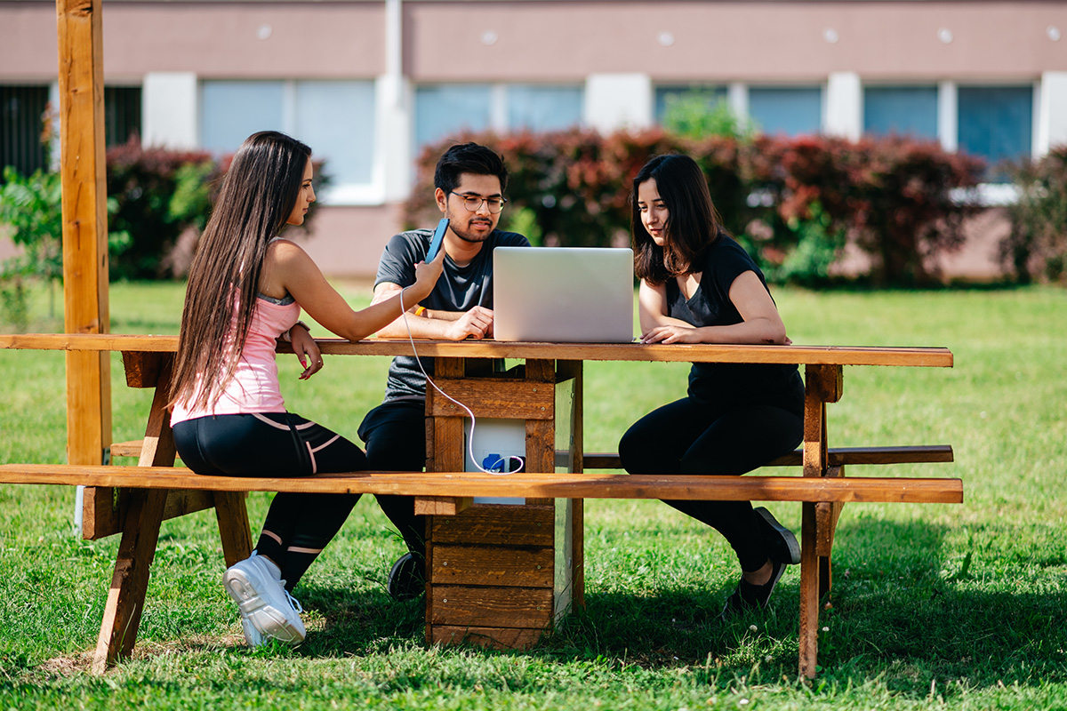 Three young people outside gathered around a table and a laptop.