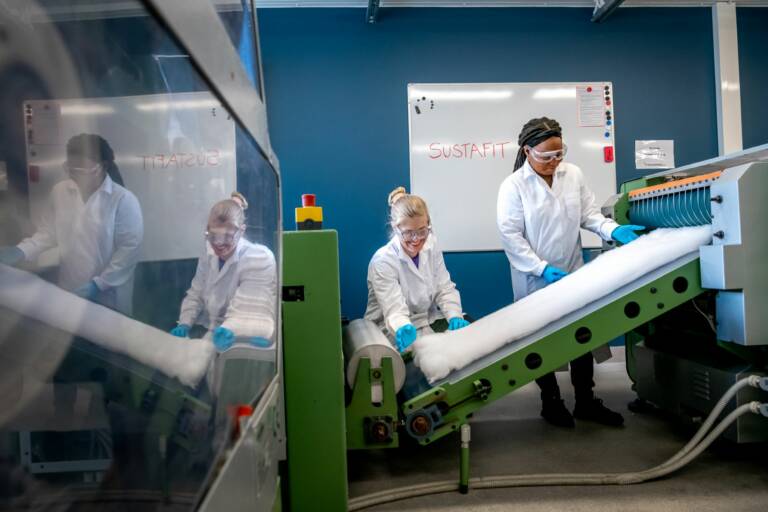 Two women working in a textile laboratory wearing white outfits and protective glasses.